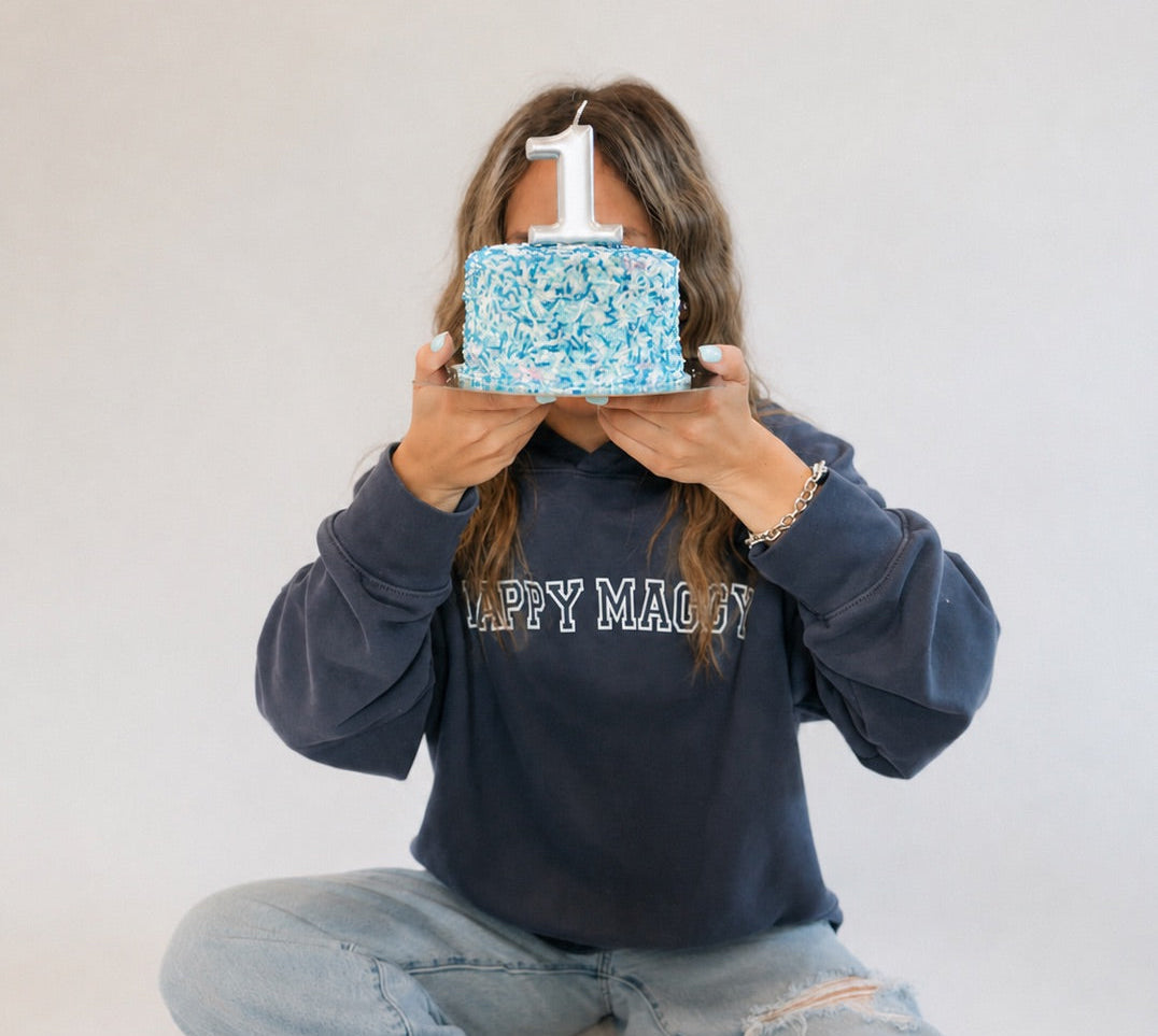 Person wearing a navy sweatshirt with 'Happy Magic' text, holding a small blue cake with white frosting.
